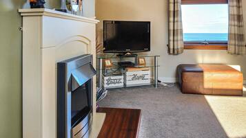 A living room with a fireplace, a television, a glass coffee table, and a view of the sea through a window.