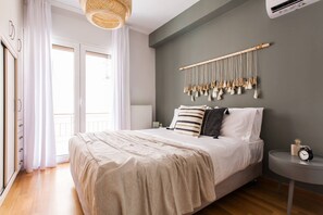 Une chambre à coucher avec un lit, une table de chevet, une décoration murale en macramé suspendue, et une vue sur l’extérieur par une fenêtre.