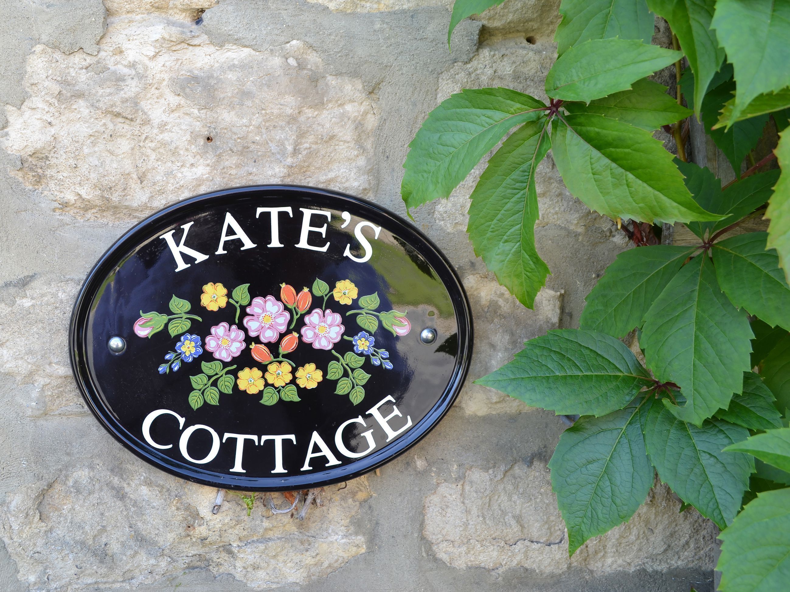 A black, oval-shaped house sign with "KATE'S COTTAGE" written in white capital letters, surrounded by colorful floral designs.