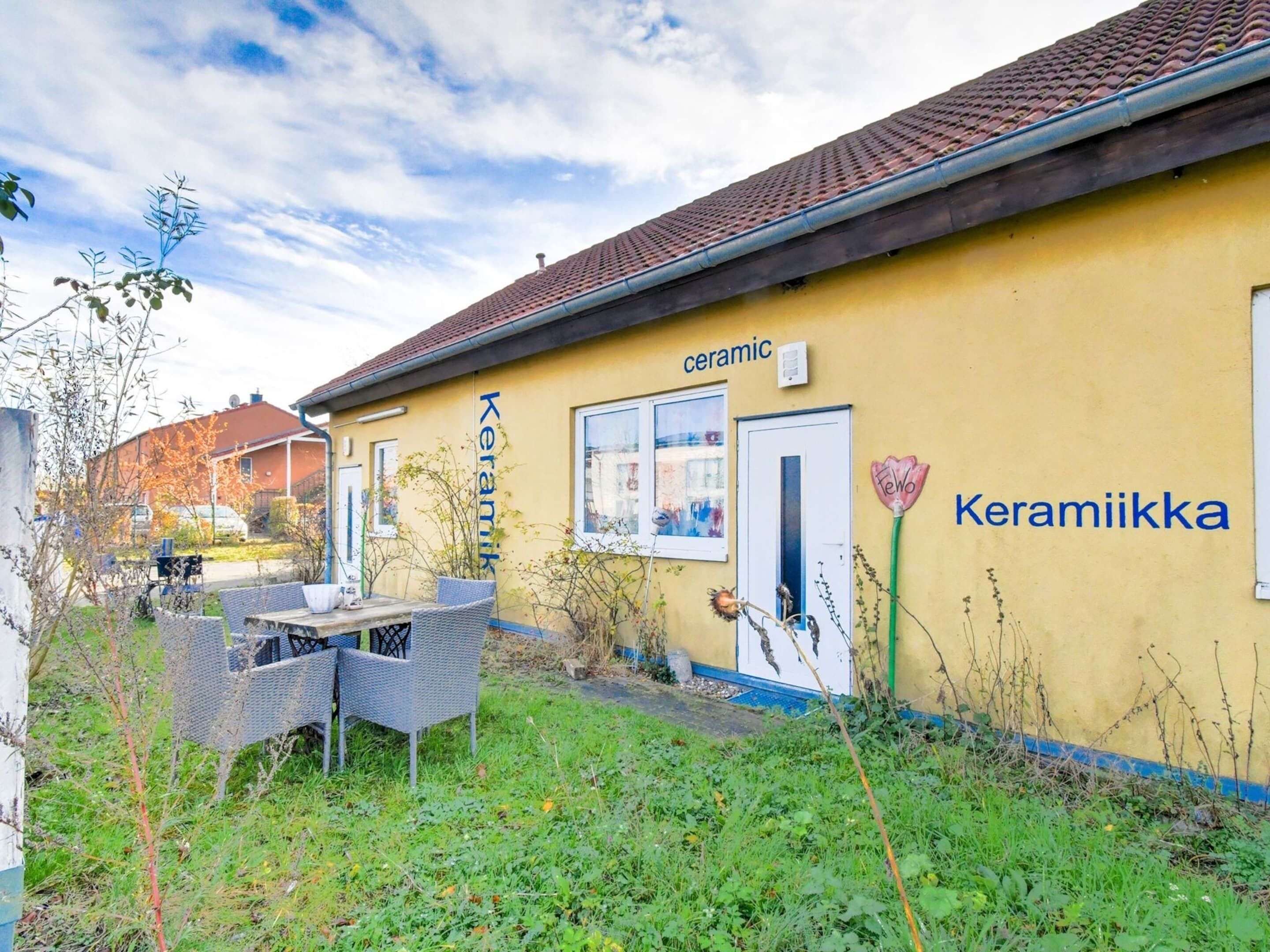 A yellow building with the words "Keramik" and "Keramiikka" on the side, a small outdoor seating area with wicker furniture, and a garden with various plants.