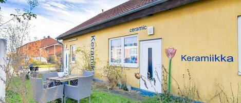 A yellow building with the words "Keramik" and "Keramiikka" on the side, a small outdoor seating area with wicker furniture, and a garden with various plants.