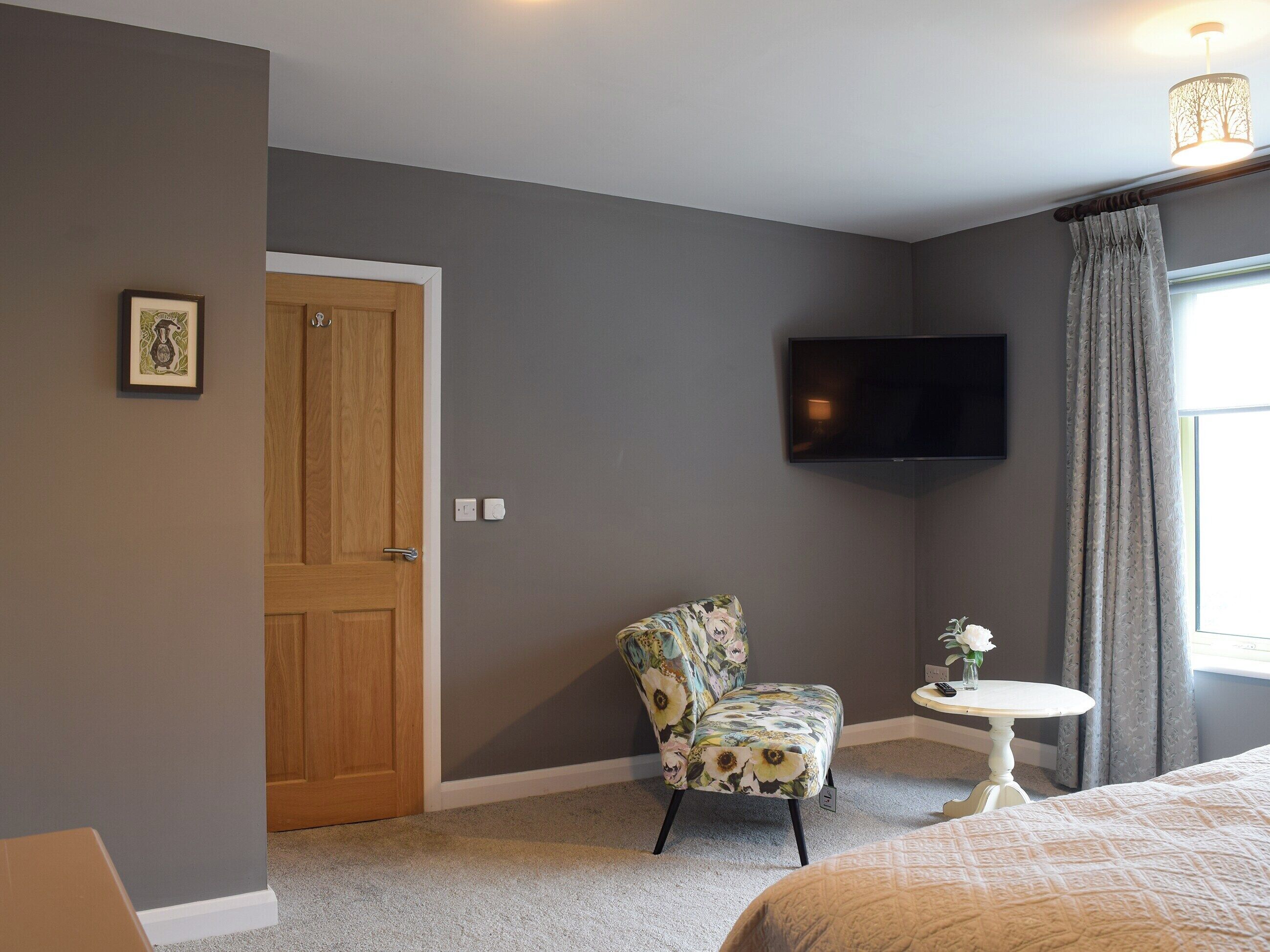 Un dormitorio con puerta de madera, un sillón floral, una mesita redonda con un florero y una cama con edredón estampado.