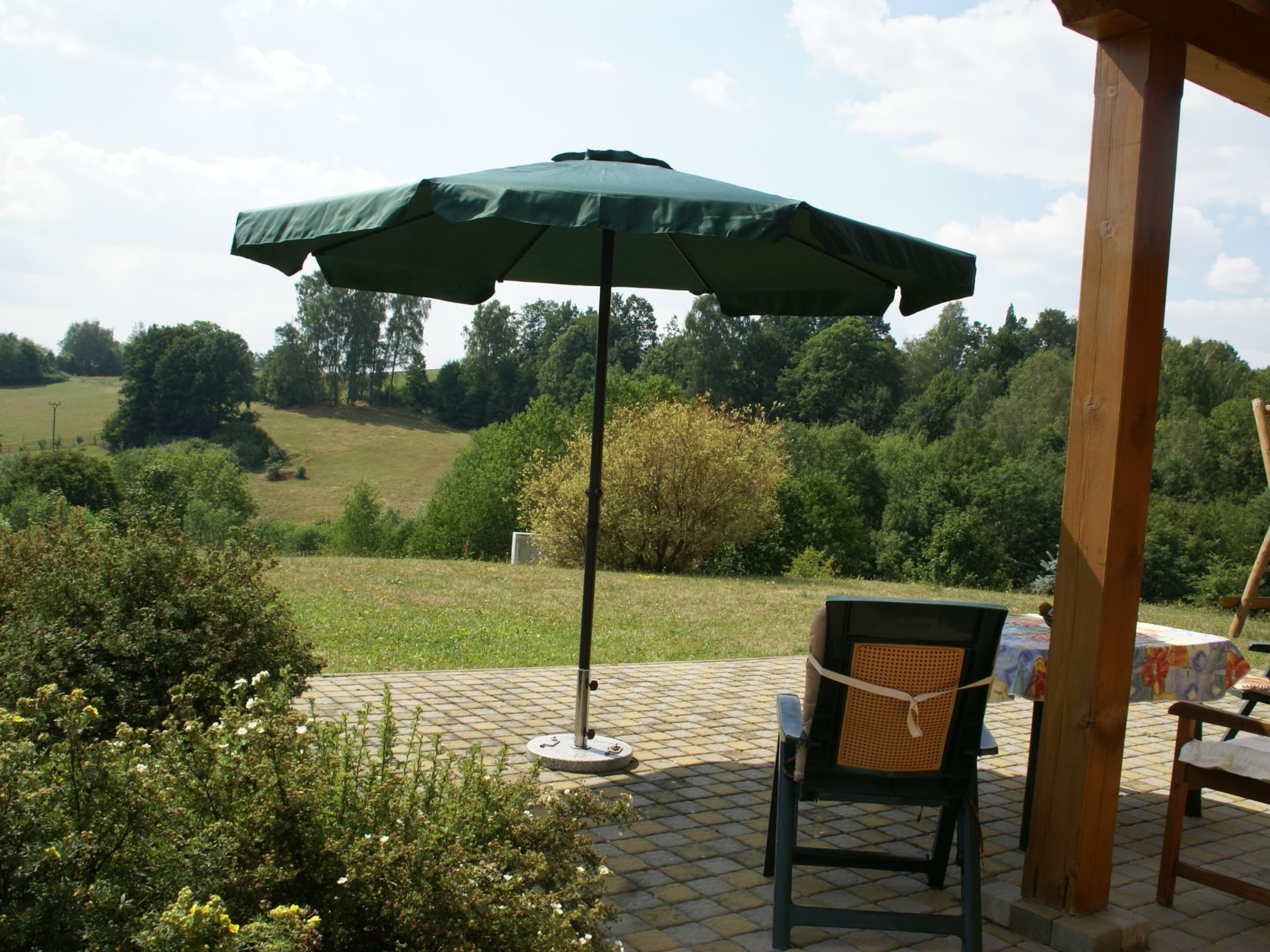 A patio with a green umbrella, a wicker chair, and a table with a floral tablecloth.