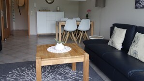 Un salon moderne avec une table basse en bois, un canapé noir agrémenté de coussins à motifs et un coin repas comprenant un buffet blanc et des chaises.