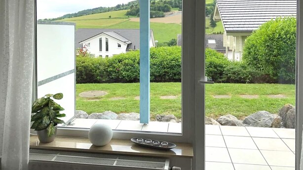 Une pièce dotée d’une grande fenêtre donnant sur un paysage verdoyant, avec des maisons et un ciel dégagé.