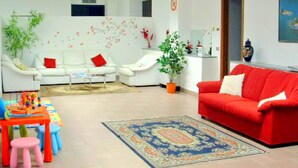 A living room with a red sofa, white armchairs, a coffee table, and a colorful rug. There are two potted plants, a small table with toys, and a red and white wall decoration. - Apartment in Residence on Lake Maggiore near Beach (Verbania)