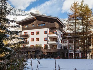 Um hotel com várias andares e varandas, chão coberto de neve e montanhas ao fundo.