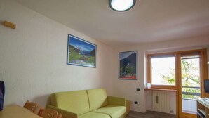 A room with a yellow sofa, wooden dining table, and chairs. There are two framed pictures on the wall, a ceiling light, and a window with a view of trees. - Matterhorn View Apartment in Breuil-Cervinia near Ski Area (Valtournenche)