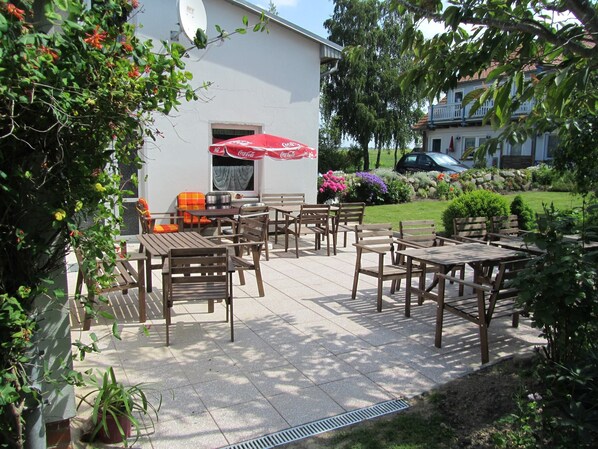 An outdoor patio area with wooden tables and chairs, a red umbrella, and a white building with a satellite dish.