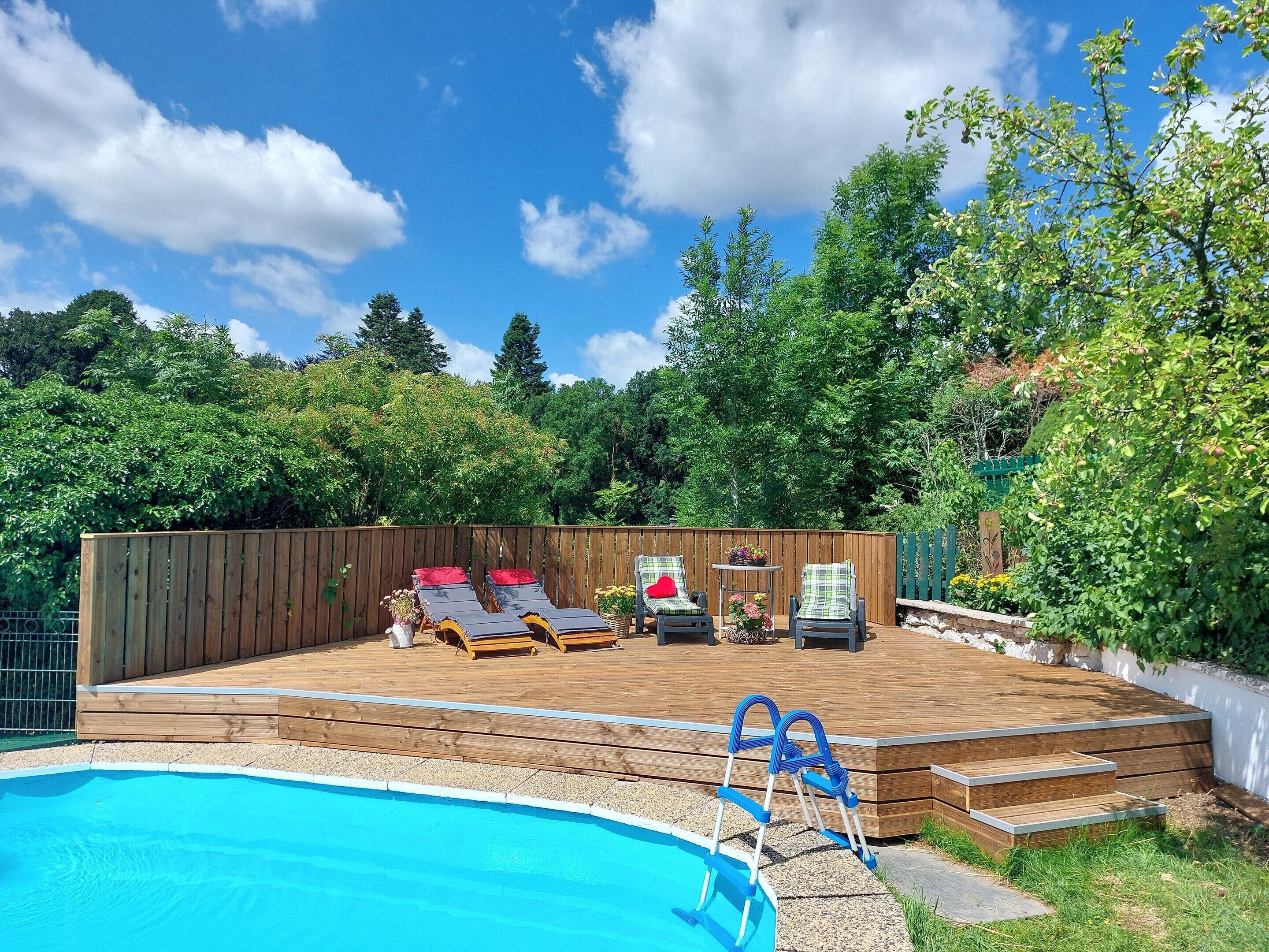 A backyard with a swimming pool, wooden deck, lounge chairs, and a wooden fence.