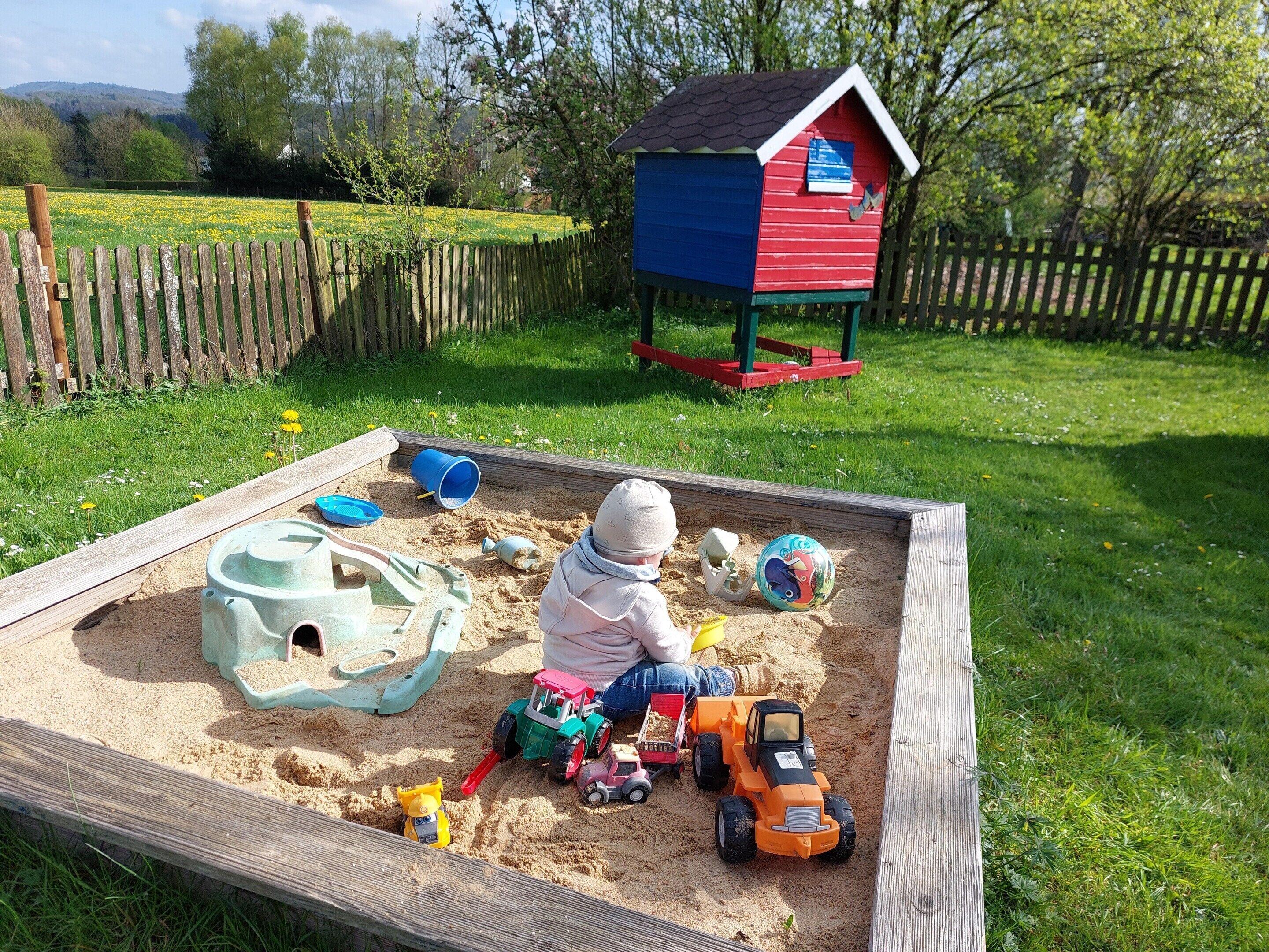 A child playing with sand toys in a sandbox with a wooden fence and a colorful playhouse in the background.