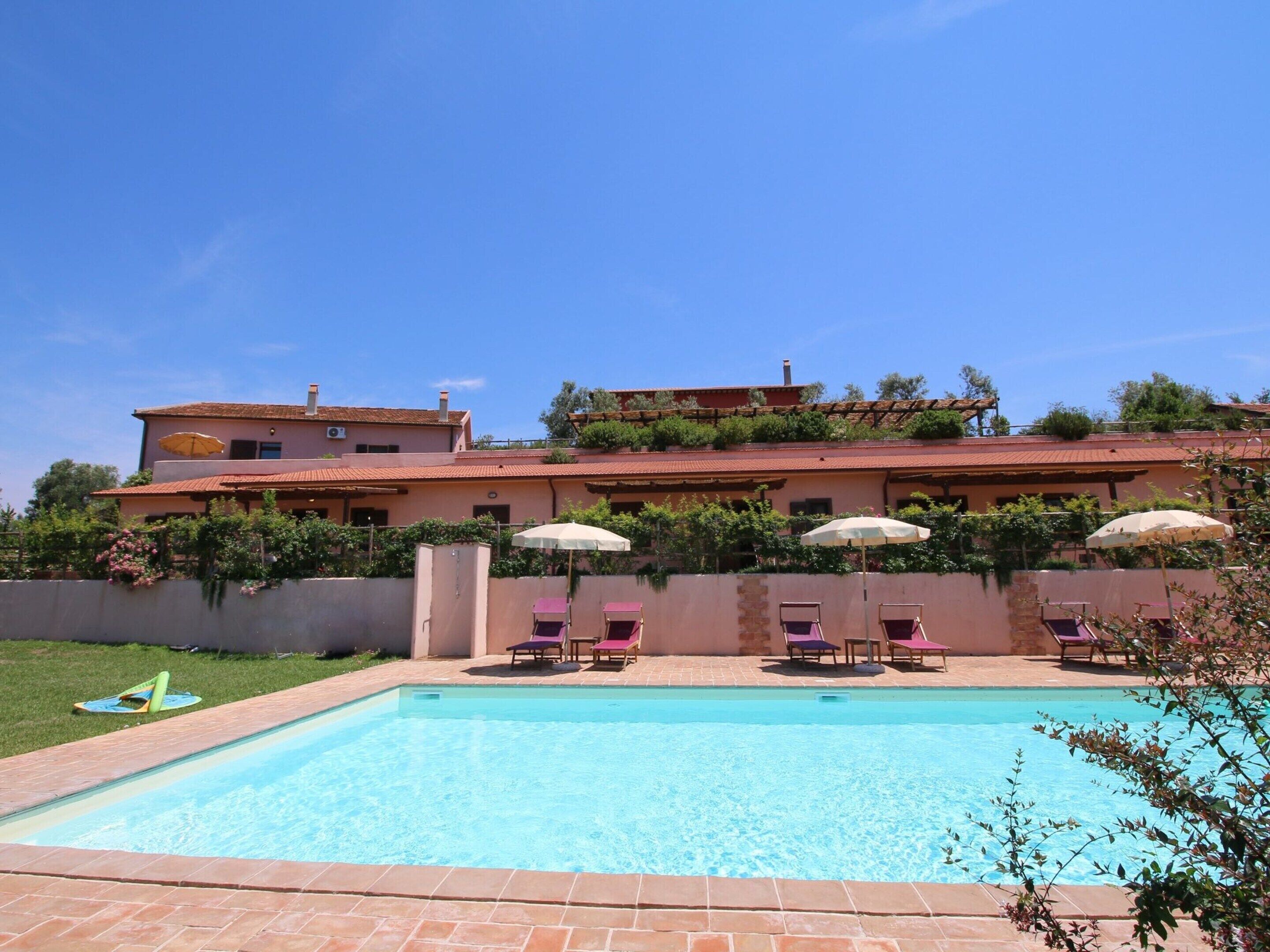 A swimming pool with lounge chairs and umbrellas, surrounded by a landscaped garden and a large building with multiple stories and balconies.