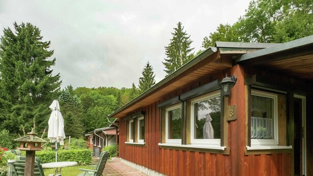 A wooden cabin with large windows, a patio with chairs and a table, and a bird feeder. - Bungalow in Neustadt Nahe Barbarossa Hohle (Harztor)