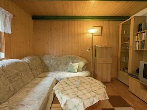 A cozy living room with a wooden floor, a sofa, a small table with a checkered tablecloth, a lamp, a cabinet with glass doors, and a television.