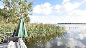 A lakeside patio with a green umbrella, wicker furniture, and a lantern. - Bootshaus Direkt am See mit Eigenem Steg an der Ostsee (Sternberg)