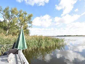 A lakeside patio with a green umbrella, wicker furniture, and a lantern. - Bootshaus Direkt am See mit Eigenem Steg an der Ostsee (Sternberg)