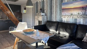 A modern living room with a black leather sofa, a wooden dining table with a bowl and a white pitcher, and a rustic wall with a sunset painting. - Bootshaus Direkt am See mit Eigenem Steg an der Ostsee (Sternberg)
