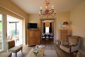 Een woonkamer met een kroonluchter, houten meubels, een televisie en een eethoek op de achtergrond.