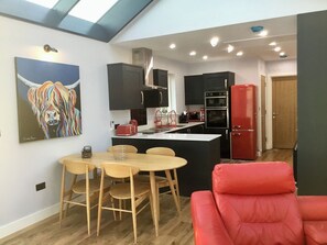 A modern kitchen with a central island, black cabinets, and a red refrigerator. There is a dining area with a wooden table and chairs, and a red sofa in the foreground. - Tidereach - Otters View - UK12269 (Connel, near Oban)
