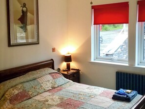 A bedroom with a bed, a nightstand with a lamp, a window with a red blind, and a radiator. - Tidereach - Otters View - UK12269 (Connel, near Oban)
