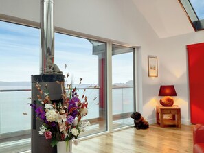 A spacious room with a large window offering a view of the sea, a fireplace with a decorative vase, and a wooden floor. - Tidereach - Otters View - UK12269 (Connel, near Oban)