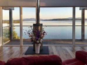 A modern living room with a large window overlooking a lake and mountains. A decorative vase with flowers is placed on a black mat in the center. - Tidereach - Otters View - UK12269 (Connel, near Oban)