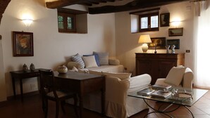 A cozy living room with a sofa, a glass coffee table, a wooden dining table, and a small kitchen area with a lamp and cabinets. - Cozy Holiday House With Terrace (Castiglion Fiorentino)