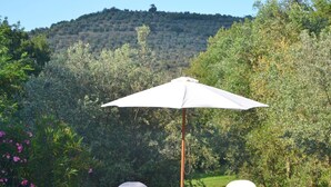 An outdoor seating area with two white chairs and a table under a white umbrella, surrounded by lush greenery and a hill in the background. - Cozy Holiday House With Terrace (Castiglion Fiorentino)