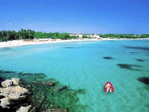 Ein Strand mit klarem türkisblauem Wasser, einer felsigen Küstenlinie und einer fernen Uferlinie mit Gebäuden.