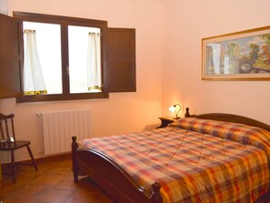 Een slaapkamer met een bed, een stoel, een radiator, een raam met gordijnen en een schilderij aan de muur.