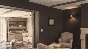 A room with a fireplace, a sofa, a chair, and a kitchen area visible through an open doorway. - Cottage in Haut-bois With Private Pool (Gesves)