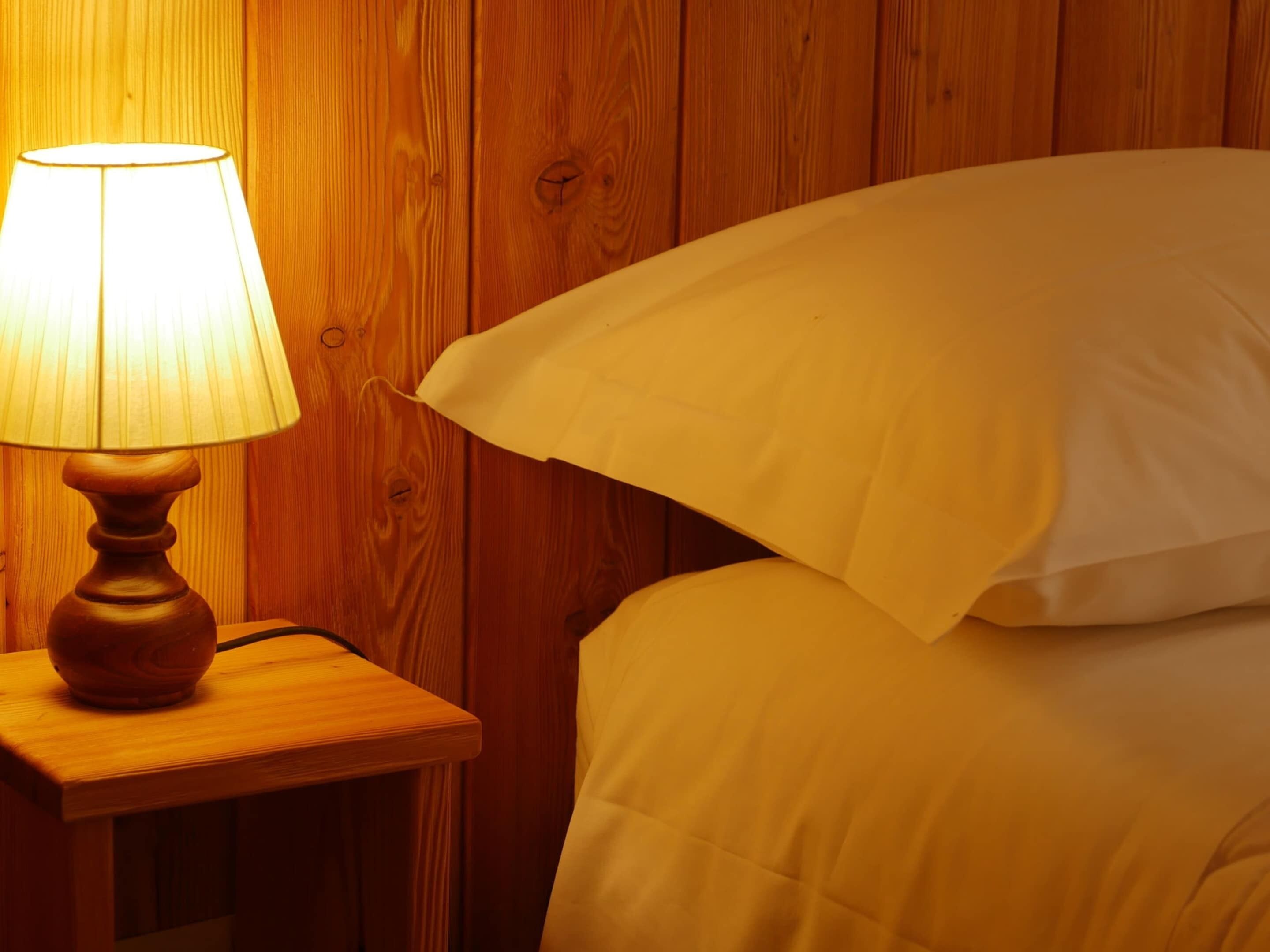 Una mesita de noche de madera con una lámpara y una cama con ropa blanca.