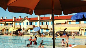 A swimming pool area with umbrellas, poolside chairs, and a building with red roofs in the background. - Sun Drenched Residence with Pool in Rosolina Mare near Venice (Rosolina)