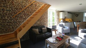 Een ruime woonkamer met een bakstenen muur, een houten trap, een zwarte leren bank, een houten salontafel en een keuken op de achtergrond.