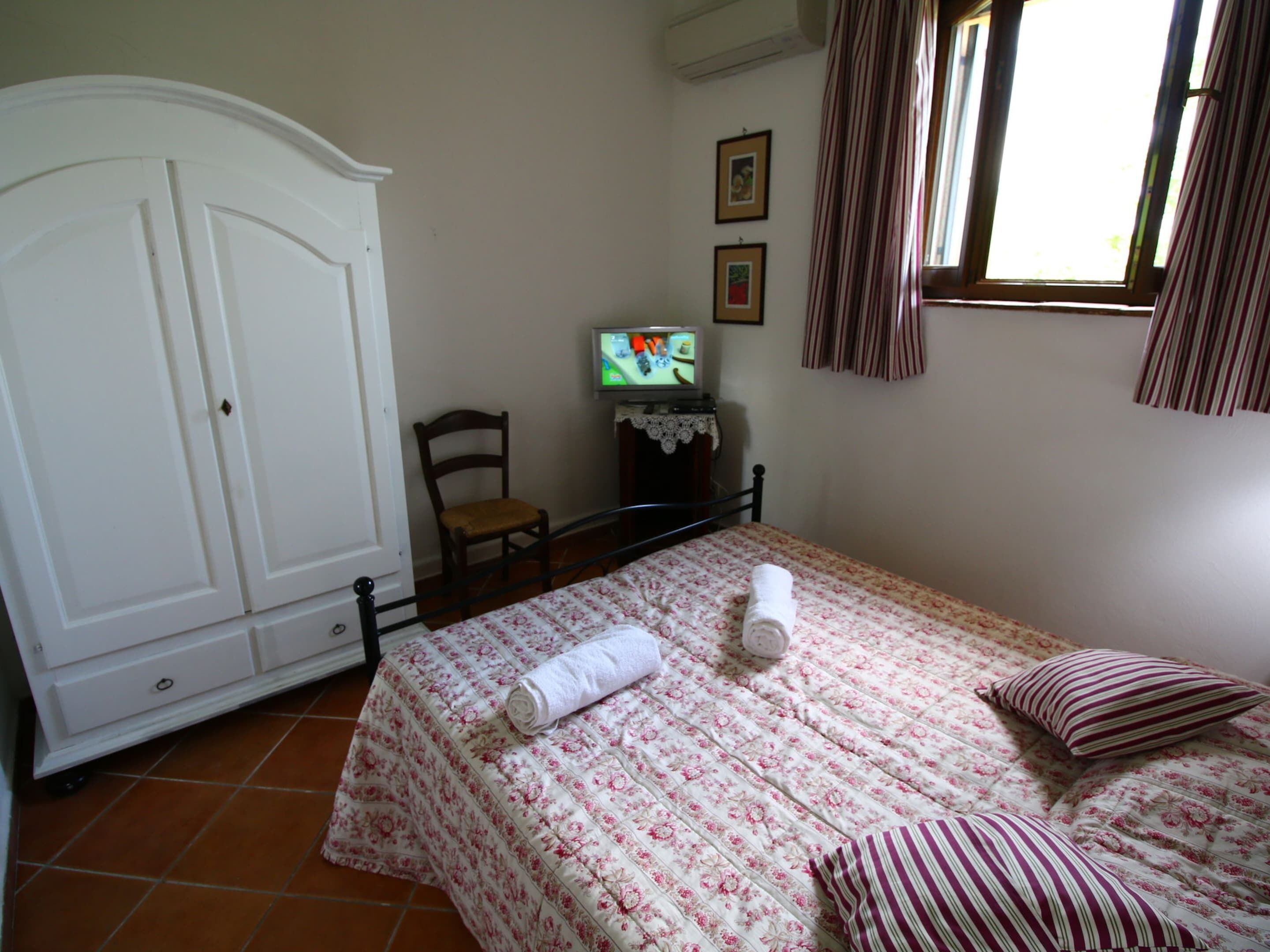 Un dormitorio con cama, una silla, un armario, televisión y ventana con cortinas.