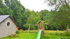 A stone cottage with a wooden playhouse and slide in a lush green garden. - Forestview Family Home (Gesves)