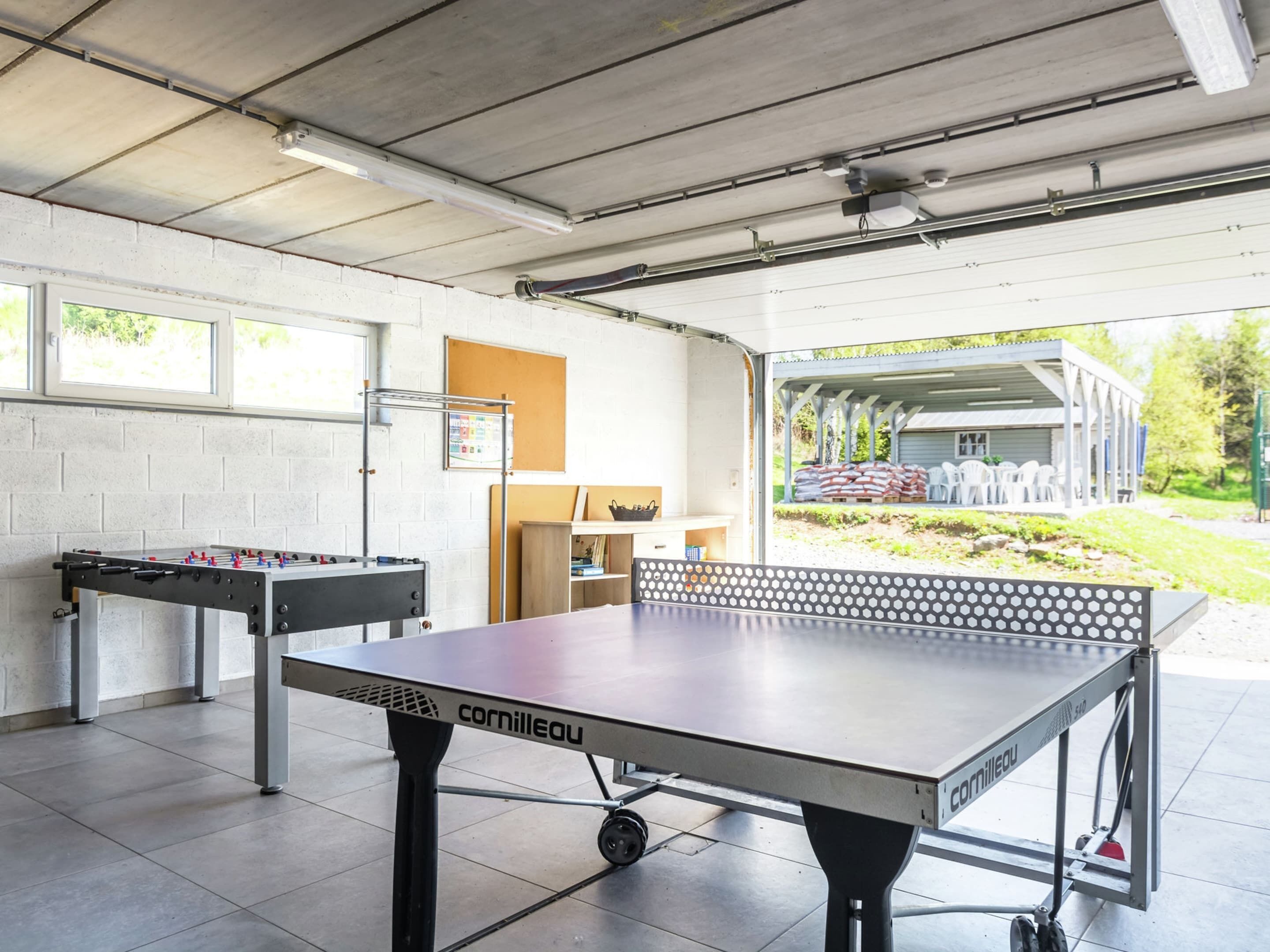 An indoor table tennis table with a visible brand name, a foosball table, and a bulletin board on a white brick wall.