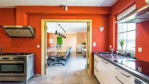 Uma cozinha moderna com fogão a gás, forno e exaustor de aço inoxidável. Há uma área de refeições com mesa e cadeiras, e uma vista para outro cômodo por uma porta aberta.