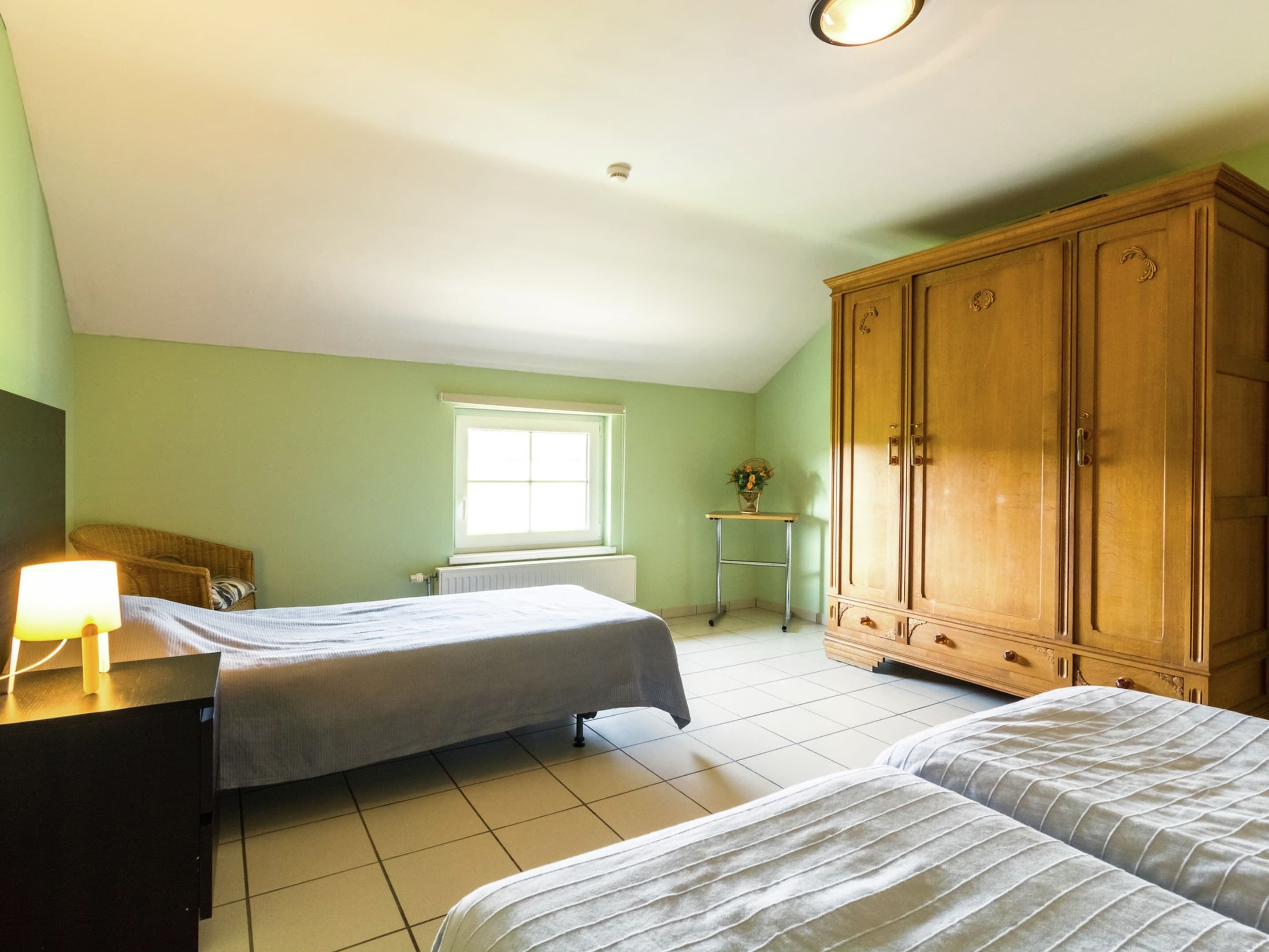 A room with two beds, a wooden wardrobe, a small table with a lamp, and a window.