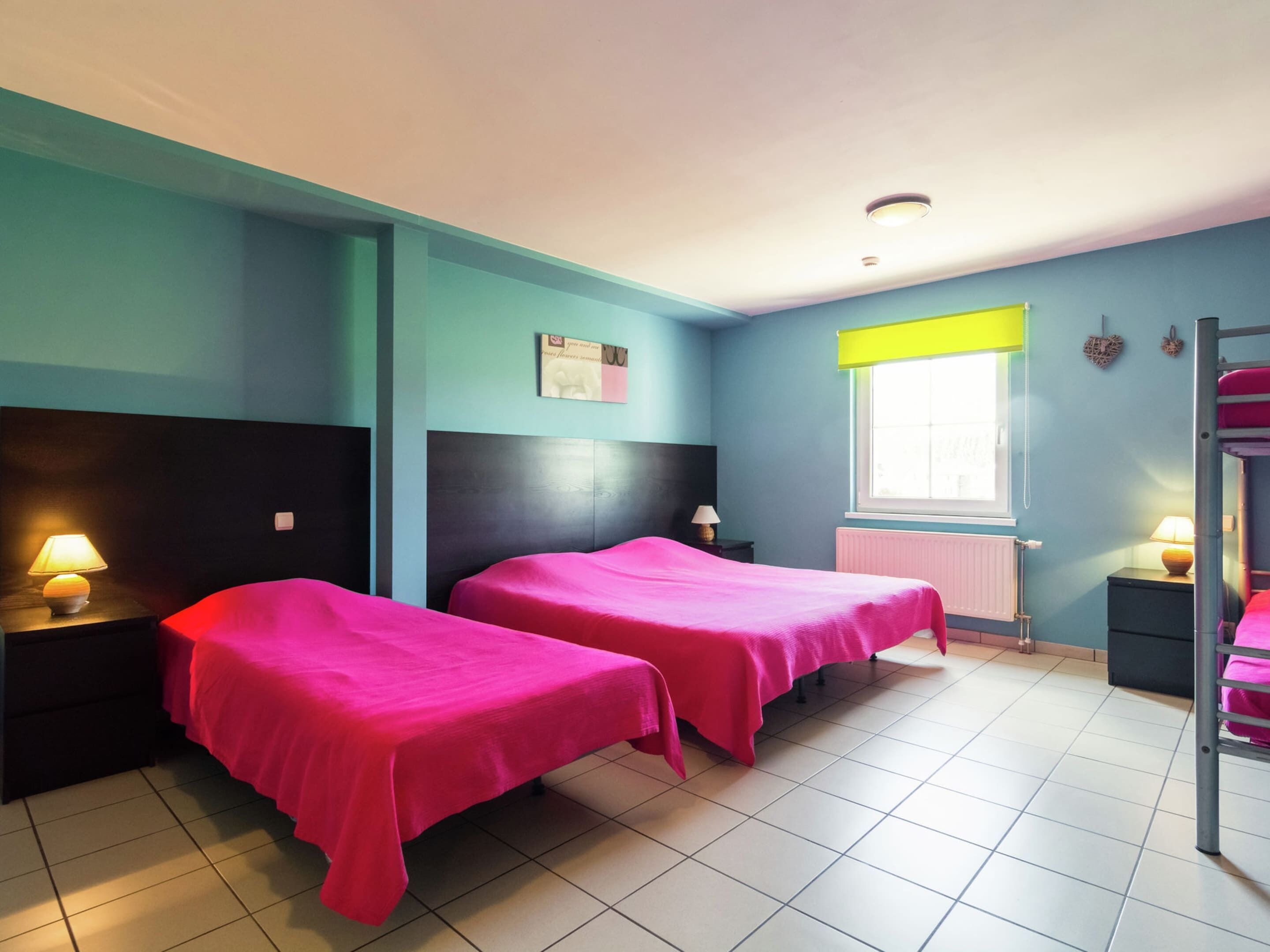 Una habitación con dos camas, una ventana con persiana amarilla, un radiador y una cama plegable en la esquina.