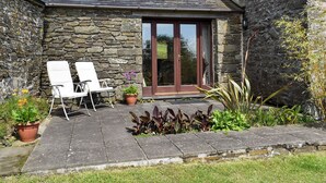 A stone building with a tiled roof, a glass door, and a small patio with two white chairs and a potted plant. - Round House - UK12083 (St Endellion, near Port Isaac)