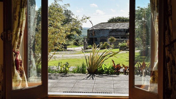 A view through an open door with curtains, revealing a stone building, a grassy area, and a clear sky. - Round House - UK12083 (St Endellion, near Port Isaac)