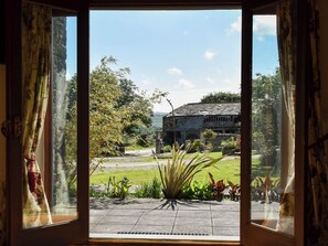 A view through an open door with curtains, revealing a stone building, a grassy area, and a clear sky. - Round House - UK12083 (St Endellion, near Port Isaac)
