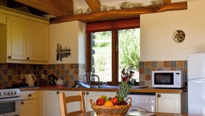 A kitchen with wooden cabinets, a microwave, a sink, and a window with a view of greenery. - Round House - UK12083 (St Endellion, near Port Isaac)