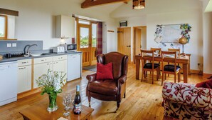 Een ruime woonkamer met houten balken, een eettafel, stoelen, een bank, een keuken met inbouwapparatuur en een uitzicht door een deur.