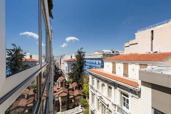 Une vue depuis une fenêtre donnant sur un paysage urbain, avec des bâtiments historiques, un ciel bleu dégagé et une étendue d’eau visible au loin.