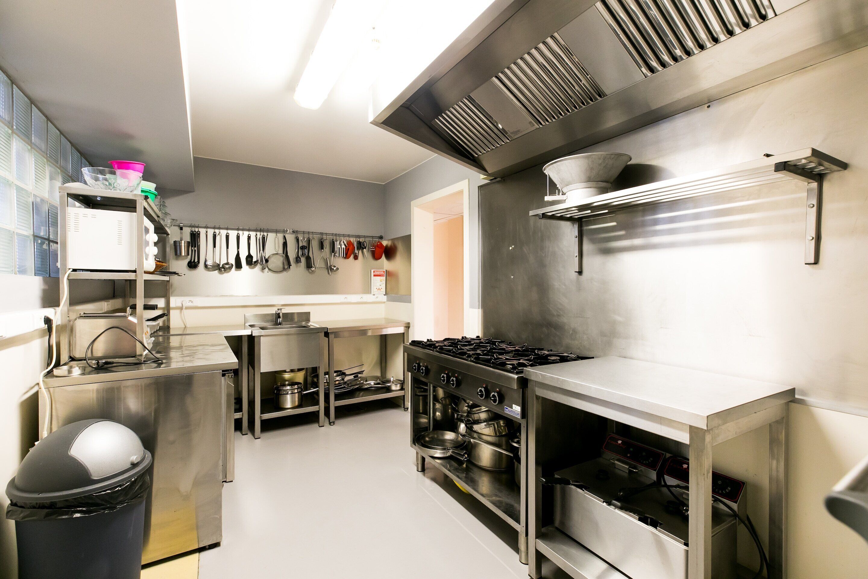 A commercial kitchen with stainless steel appliances, a pot rack, and a trash bin.
