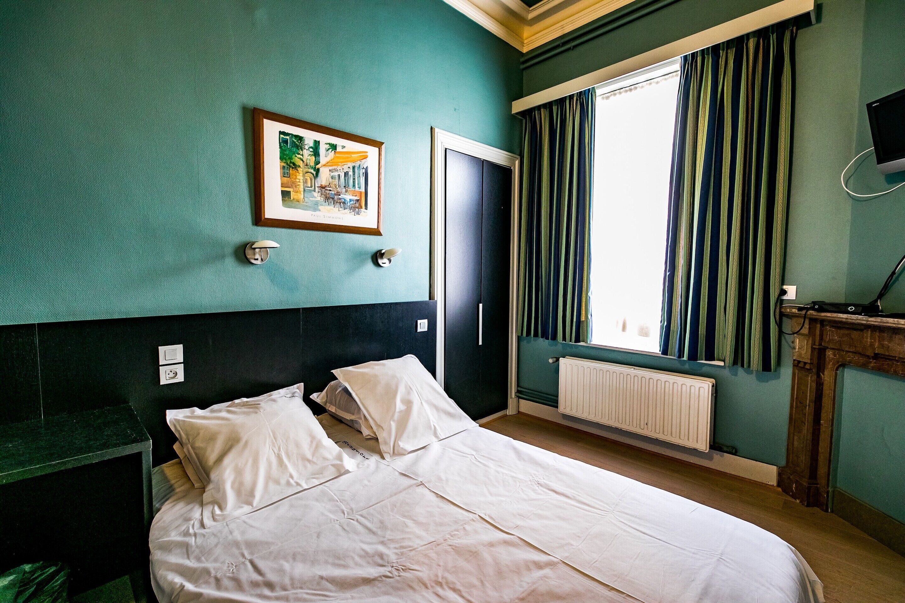 Ein Zimmer mit einem Bett, einem Bild an der Wand, einem Fenster mit Vorhängen, einem Heizkörper und einem kleinen Tisch mit einem Fernseher.