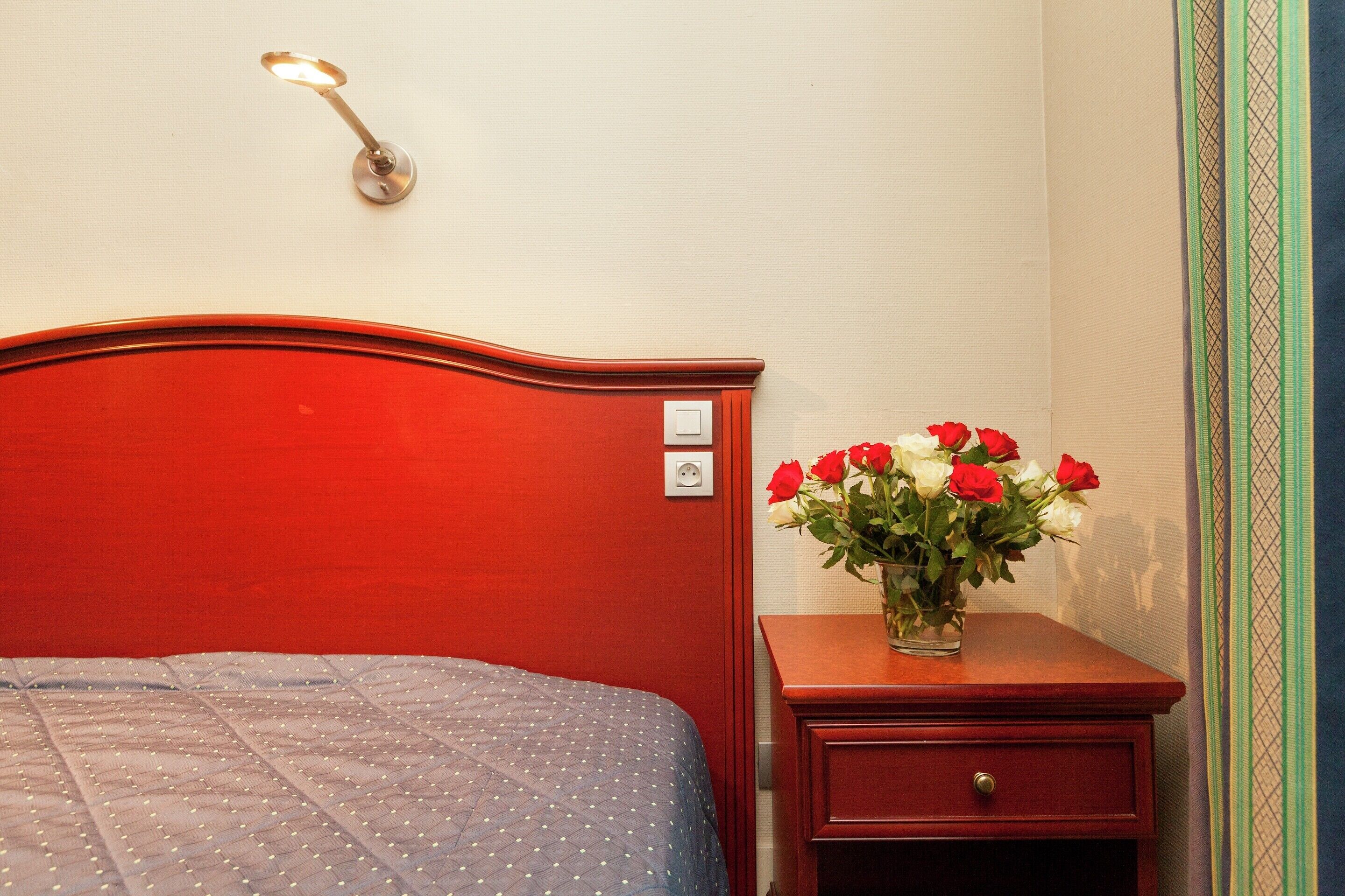 Una cama con cabecera roja, una mesita de noche con un florero de rosas rojas y blancas, y una lámpara de pared.