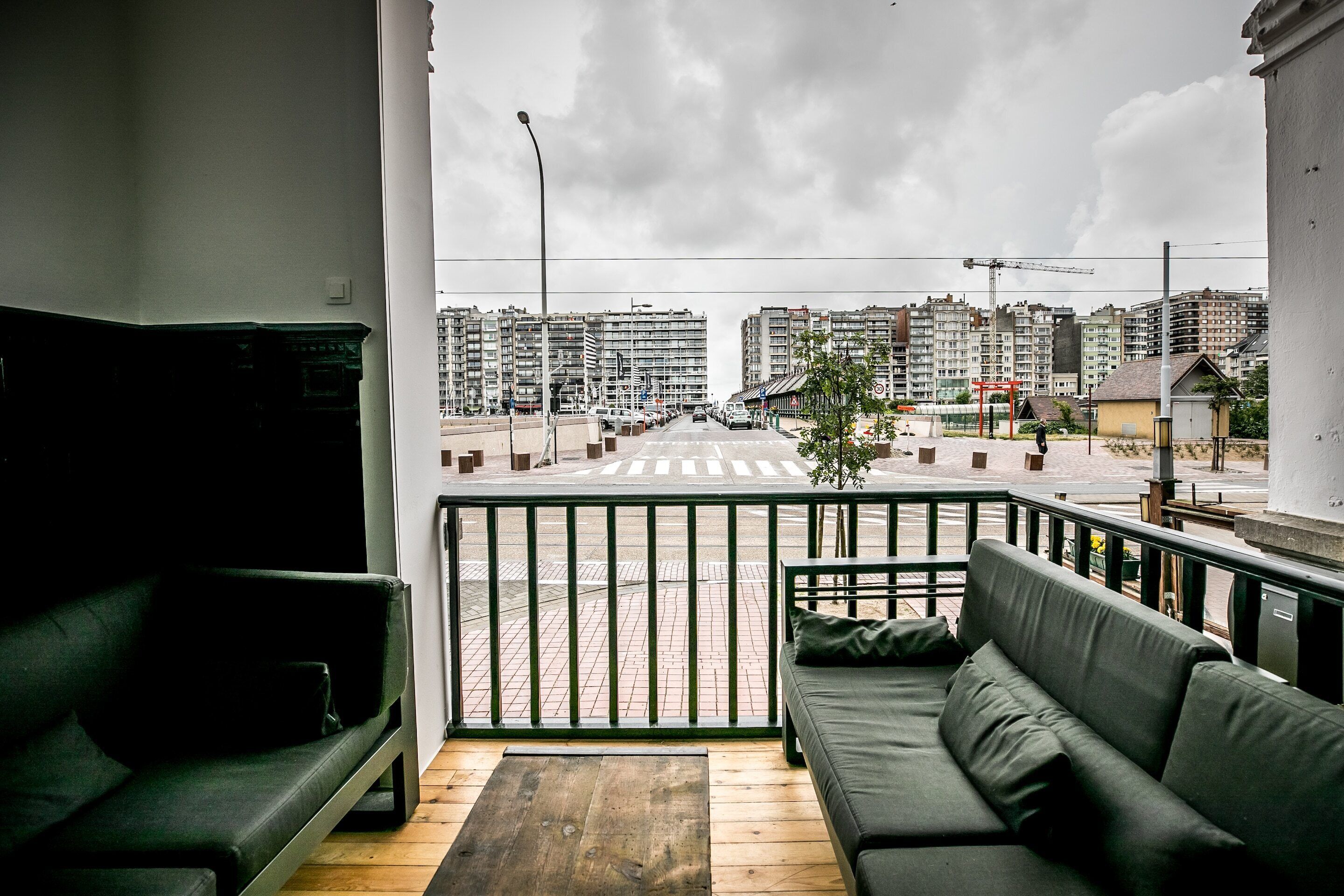 A balcony with a sofa and a wooden table, overlooking a street with buildings and a pedestrian crossing.